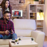 A guy is sitting on a couch with a joystick and playing a console.